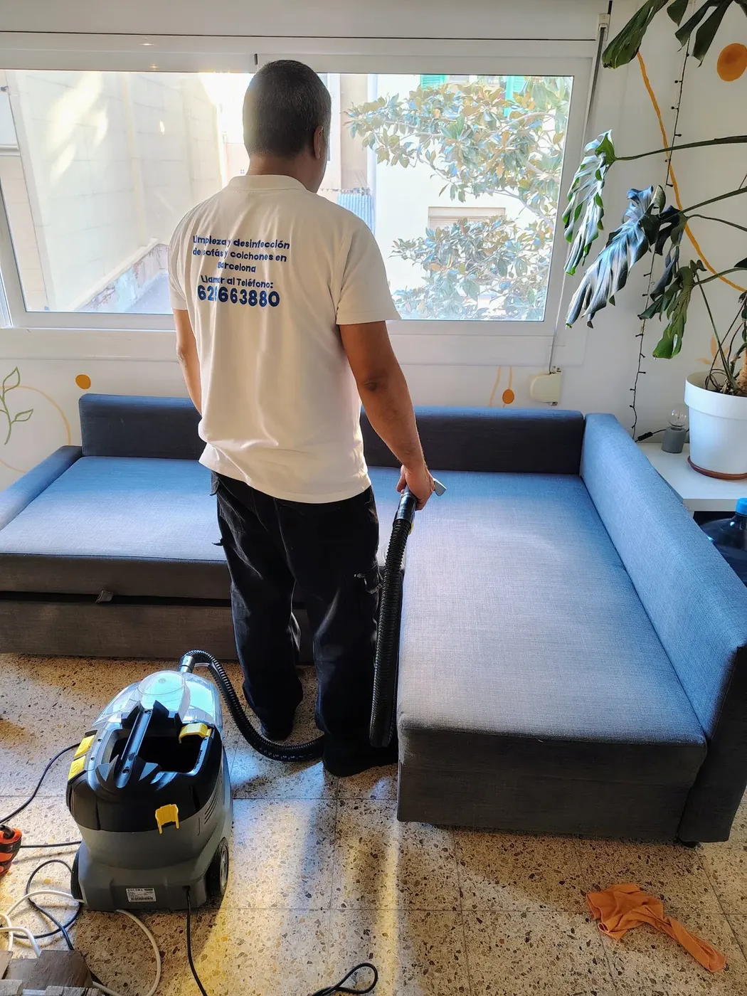 Técnico de Vaporex Barcelona, con camiseta de la empresa, limpiando un sofá azul junto a una ventana